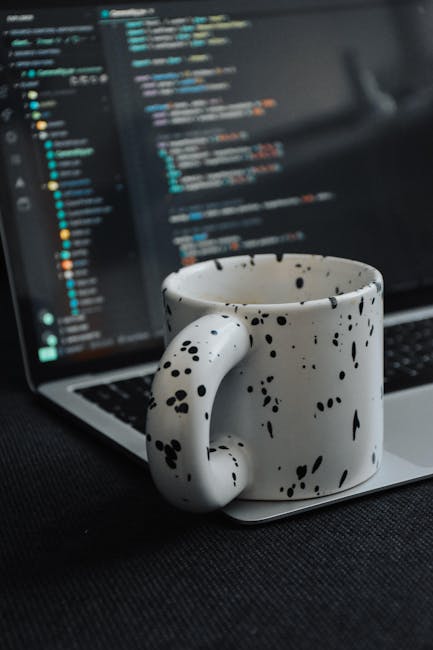 A close-up view of a white ceramic mug with black speckles sitting on a laptop keyboard, which displays lines of colorful code on its screen. Behind the mug, the laptop's screen reveals a blurred codi