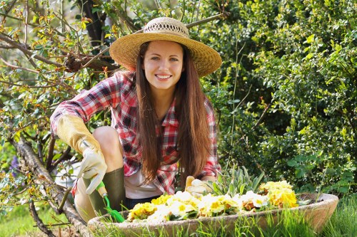 A young woman with long brown hair, wearing a straw sun hat, yellow gardening gloves, a red and white checkered shirt, and grey trousers, is kneeling in a lush garden during a sunny day. She is smiling as she tends to a flower bed with vibrant yellow and purple flowers, which is bordered by a wooden edge. Behind her, there is a dense backdrop of green foliage, including leafy trees and shrubs, creating a natural outdoor environment. The garden features a well-maintained grassy lawn in the foreground, with a mixture of soil and plant borders extending into the background. Sunlight filters through the leaves, illuminating the scene and highlighting the healthy, vibrant plants. The setup indicates active gardening work, suggesting outdoor maintenance and plant care services provided by Gardening Services Downham, with a natural and tidy landscaped garden scene typical of family or neighborhood outdoor spaces in the Downham area.